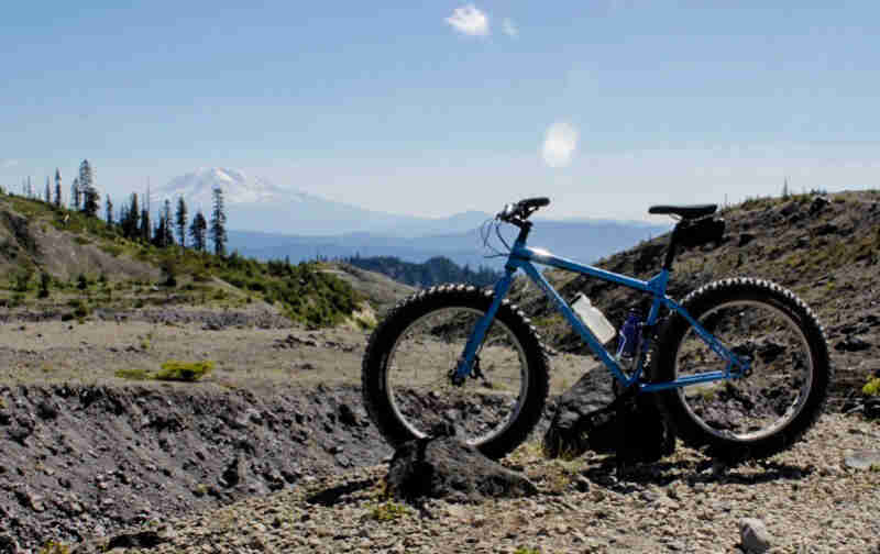 Left view of a Surly fat bike, blue, parked on a rocky field, with Mt. St. Helens in the background