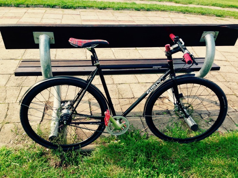 Right side view of a black Surly Cross Check bike, parked on grass along the back of a park bench on a sidewalk