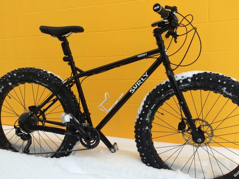 Right side view of a black Surly Pugsley fat bike, parked in snow next to a yellow, cinder block wall