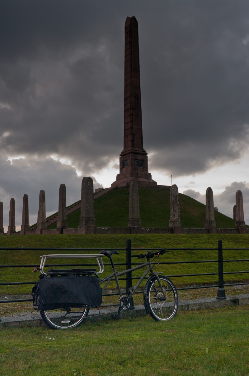 Right side view of a Surly Big Dummy bike, parked on grass against a steel fence, with a tall, stone monument behind