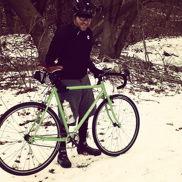 Right side view of a green Surly bike parked on the snow, with a cyclist standing behind and woods in the background