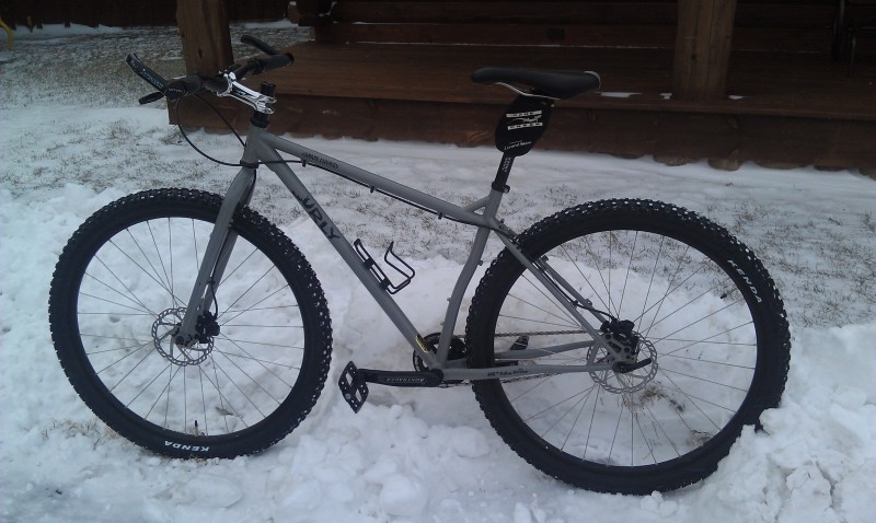 Left side view of a gray Surly Karate Monkey bike, standing on snow, in front of a wood porch