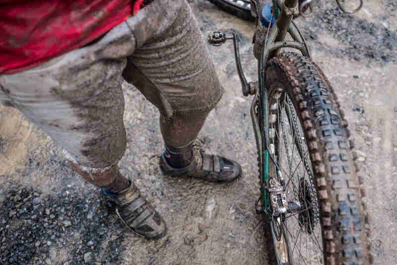 Downward cropped view of the muddy legs of a cyclist, standing next to the back wheel of their dirty bike