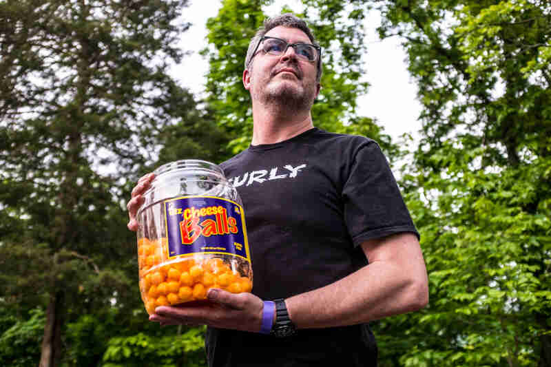 Front view of a person in a black Surly t-shirt, holding a plastic jar of cheese balls, with trees in the background