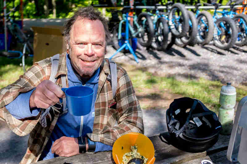 Front view of a person sitting at a picnic table, holding up a cup, with a row of blue tat bikes in the background