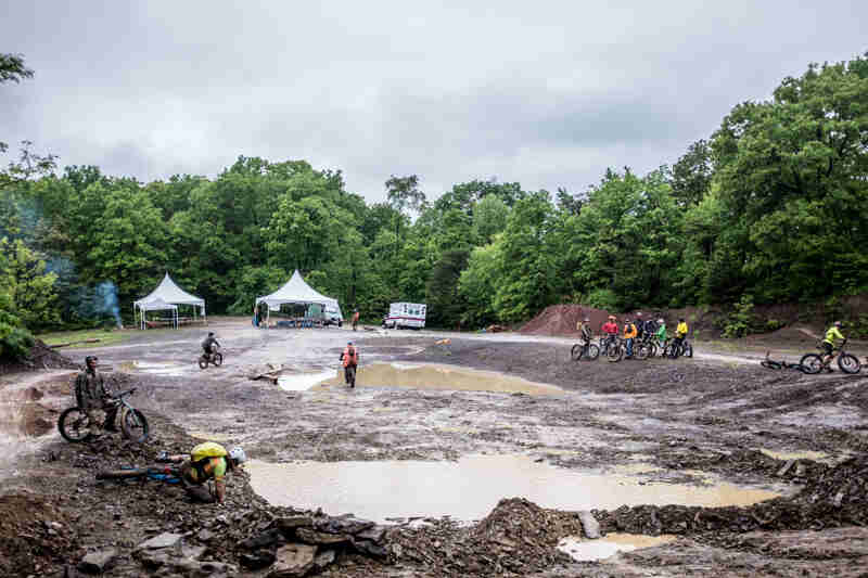 Cyclists standing around a muddy bike skills course, with canopies, an ambulance and trees in the background