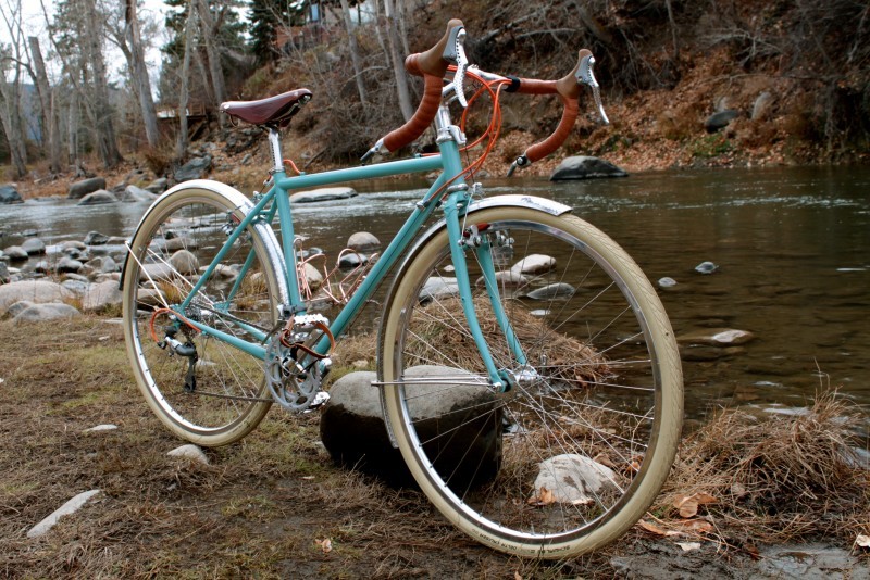 Right side view of a mint Surly Cross Check bike, on a flat bank next to a river, with bare woods in the background