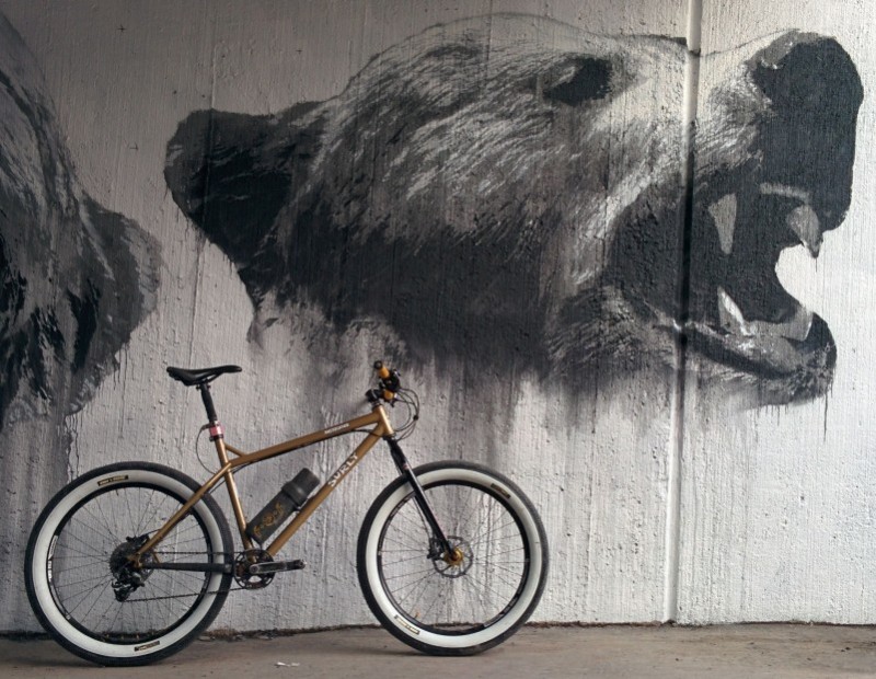 Right side view of a sand color Surly Instigator bike with white wall tires, leaning on a wall with a bear head mural