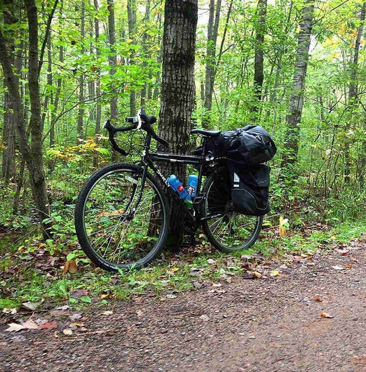 Left side view of a Surly bike, loaded with gear, on the side of a gravel road, with the woods in the background