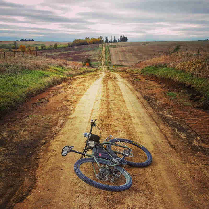 Rear view of a Surly bike, laying on it's left side, facing down a gravel country road, between farm fields