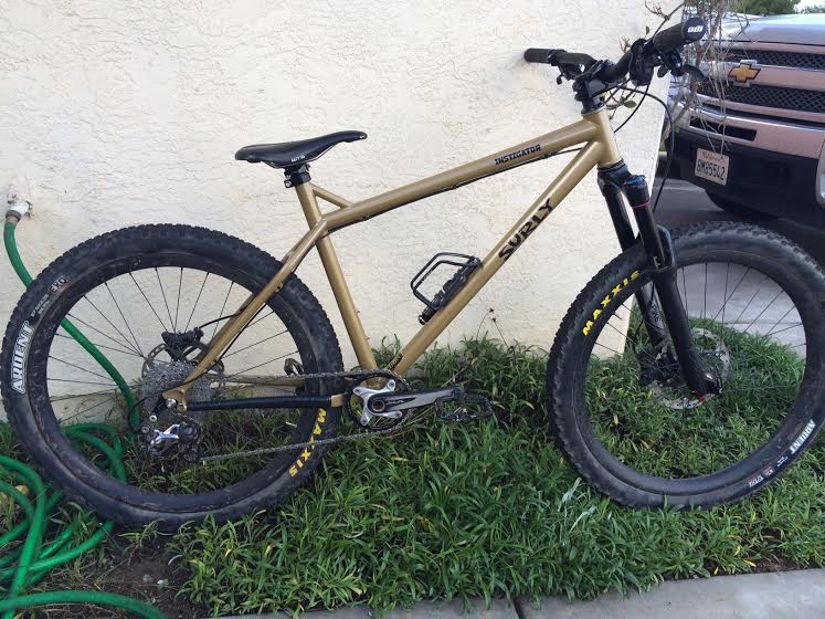 Right side view of a tan Surly Instigator bike, parked on grass between a walkway and a stucco wall
