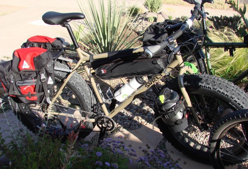 Right side view of a tan Surly fat bike with gear, parked in front of a landscaped plot, with grasses and cacti