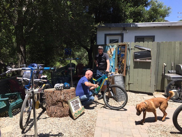 Two people repairing a bike and a dog on a walkway, in front a gate with a house behind, and trees in the background