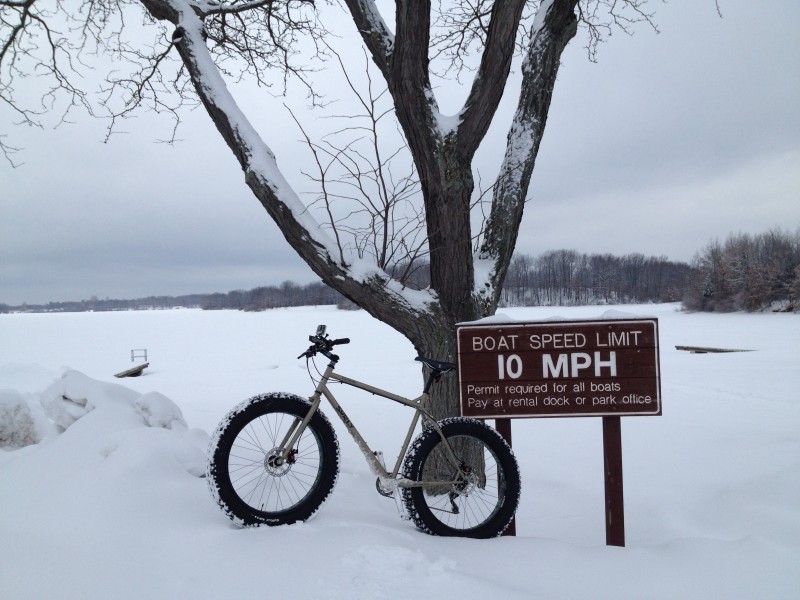 Left side view of a tan Surly Moonlander fat bike, leaning on a boat speed limit sign, on a snowy bank of a frozen lake