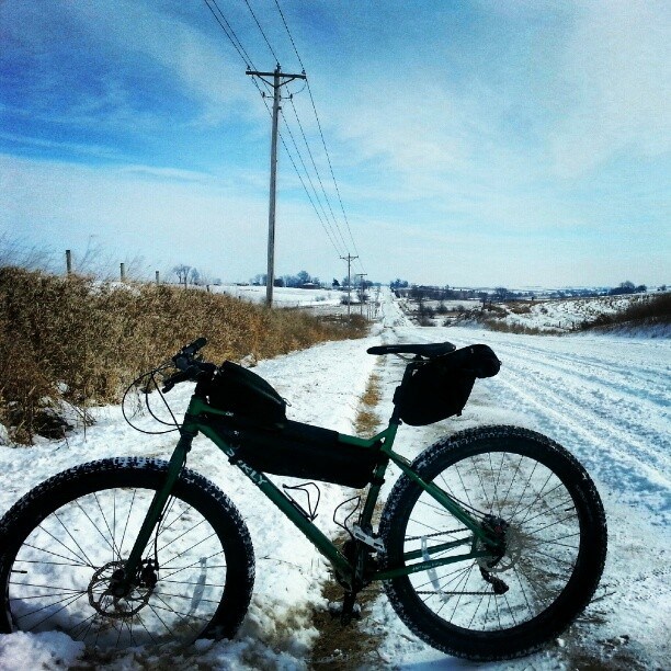 Left side view of a green Surly Krampus bike, parked across the side of a snow covered gravel road in the country