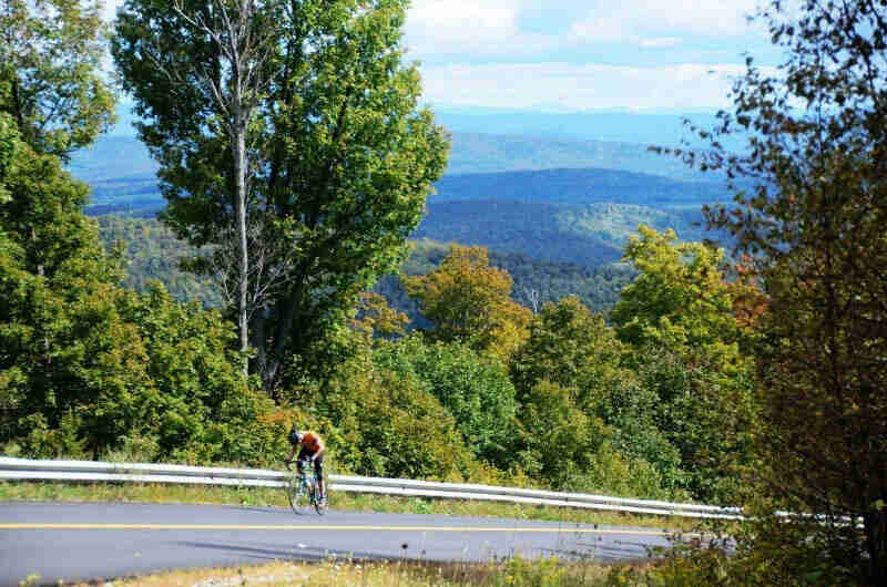 Left side of a cyclist riding up a steep hill on a paved road, with tree covered mountains in the background