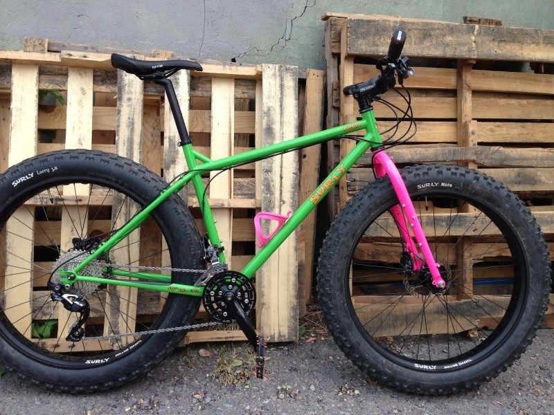 Right side view of a green Surly Pugsley fat bike with pink forks, leaning on pallets that are standing against a wall