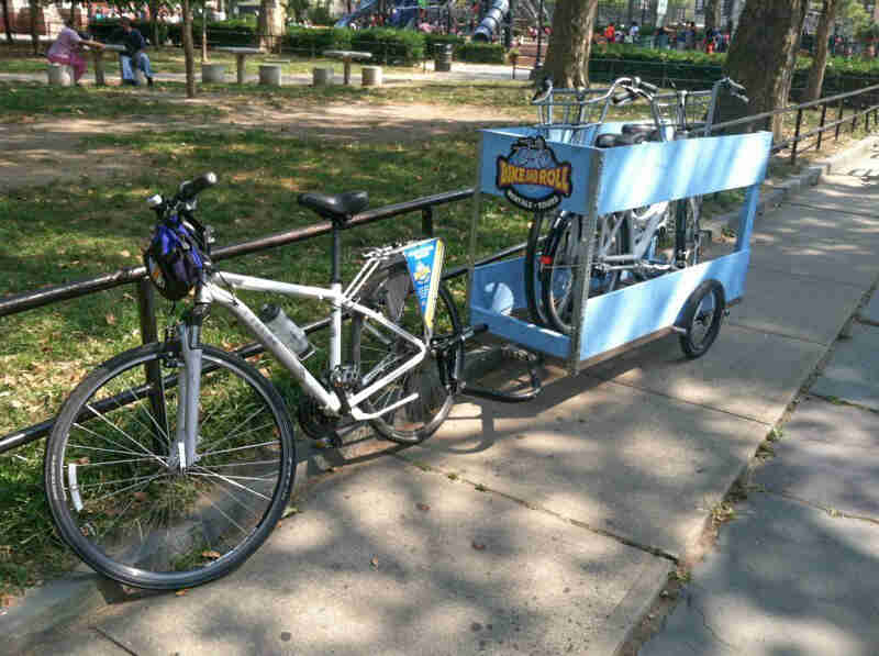 Left side view of a bike with a trailer, parked on a sidewalk, against a railing, with a park in the background