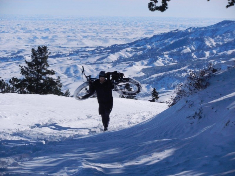 Front view of a cyclist, carrying a Surly fat bike on their back, up a snow covered hill, with snowy hills in background