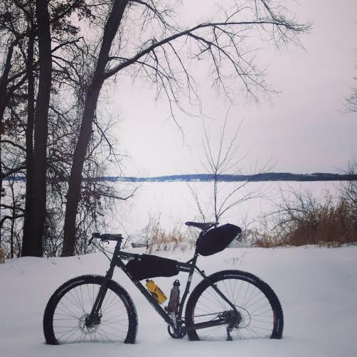 Left side view of a Surly Ogre bike with gear, parked in deep snow, with trees and a frozen lake in the background