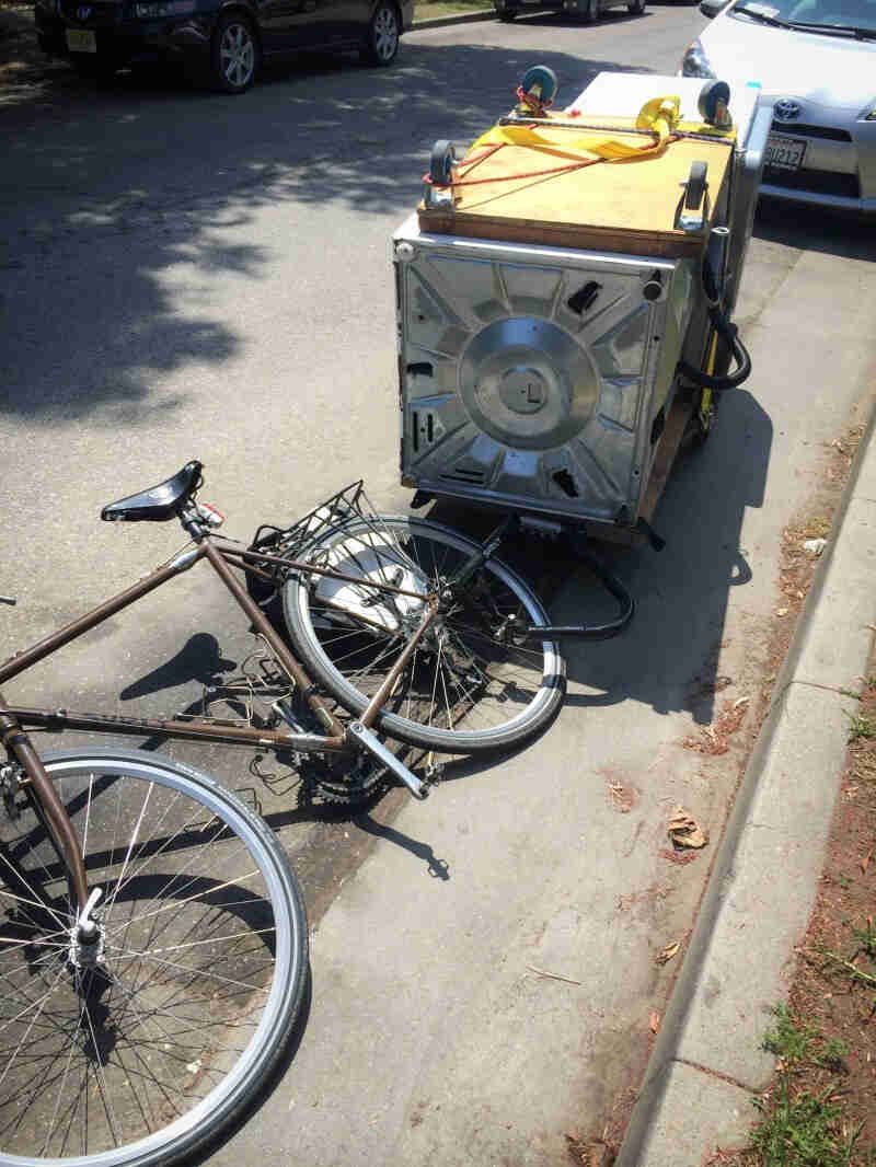 Downward, left side view of a brown Surly bike, laying on it's side, with a washer & dryer on a trailer, on a streetside