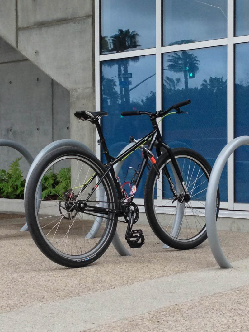 Rear, right side view of a black bike, parked against a bike rack in front of a glass wall, outside of a building 