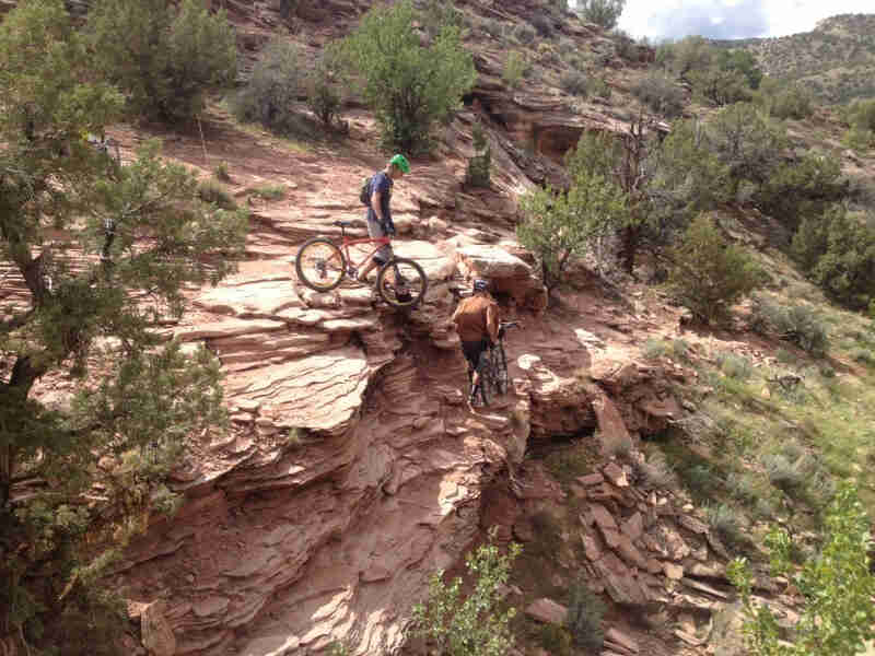 Downward view of 2 cyclists, climbing down a desert cliff wall with their Surly bikes in hand