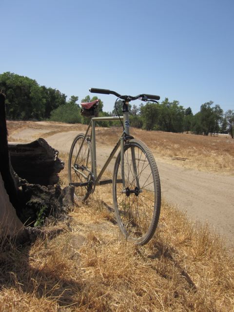 Front, right side view of a tan Surly bike, parked on brown grass beside a dirt trail, with a field in the background
