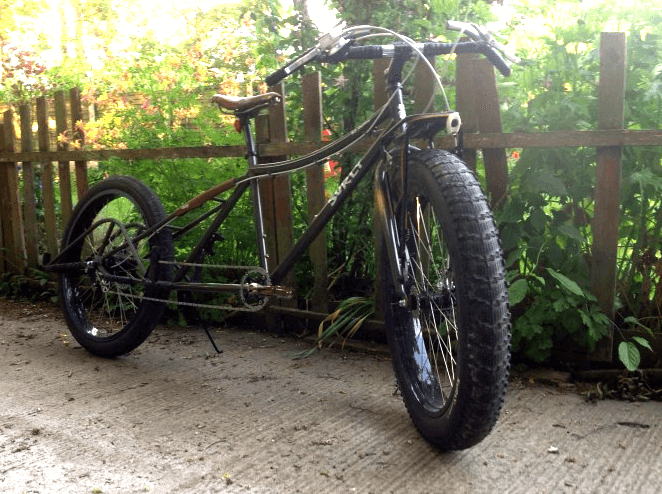 Front, right side view of a Surly Big Fat Dummy bike, leaning wood fence on a cement slab, with bushes behind it