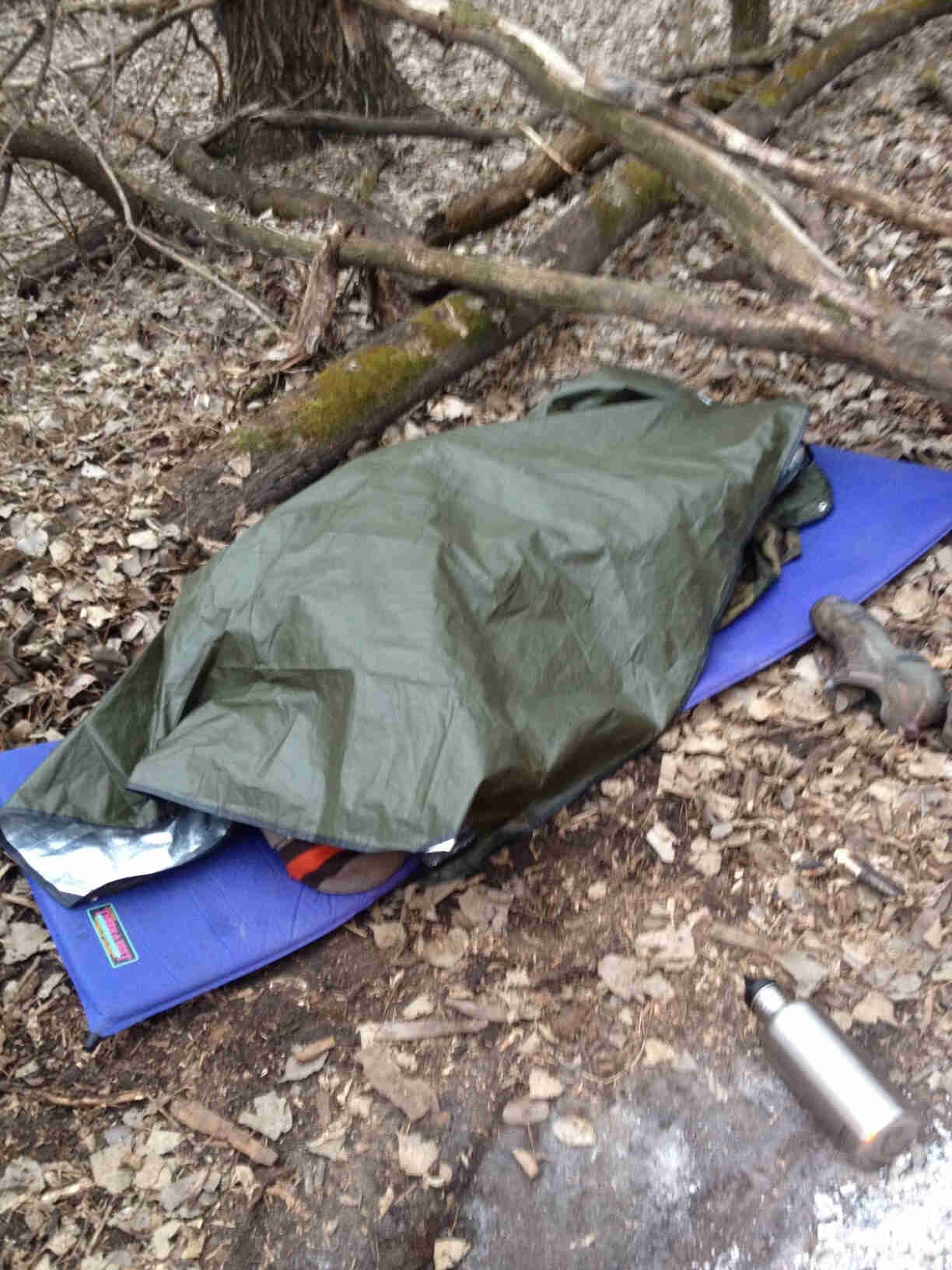 An olive color tarp covers camping gear, on top of dirt and leaves with tree branches laying beside