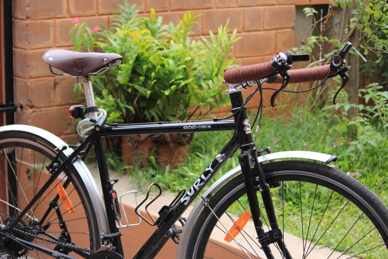 Right side view of a black Surly Cross Check bike, leaning on a short brick wall, with grass area above and behind
