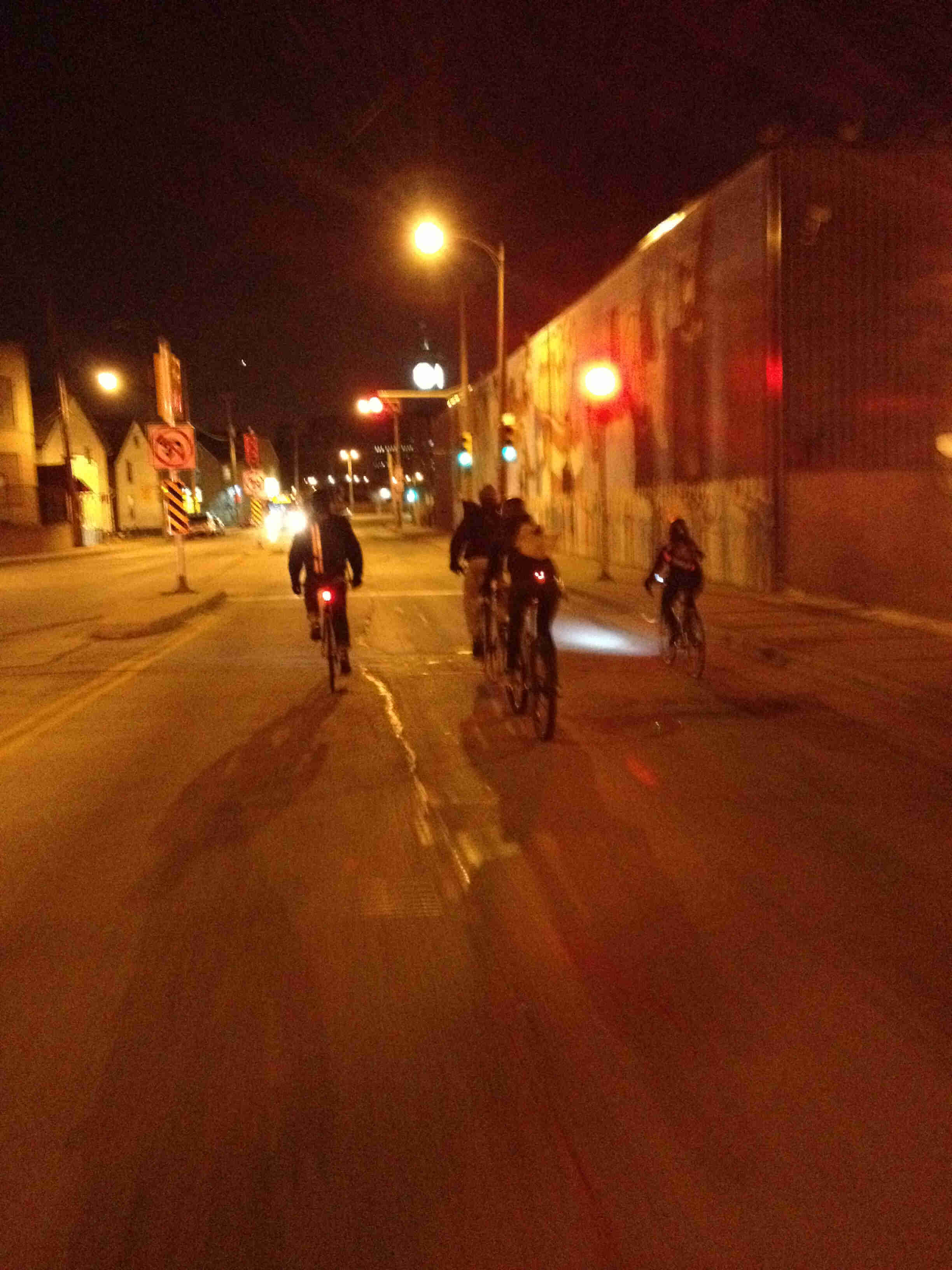 Right view of a group of cyclists riding on a street at night, with buildings on both sides of the road