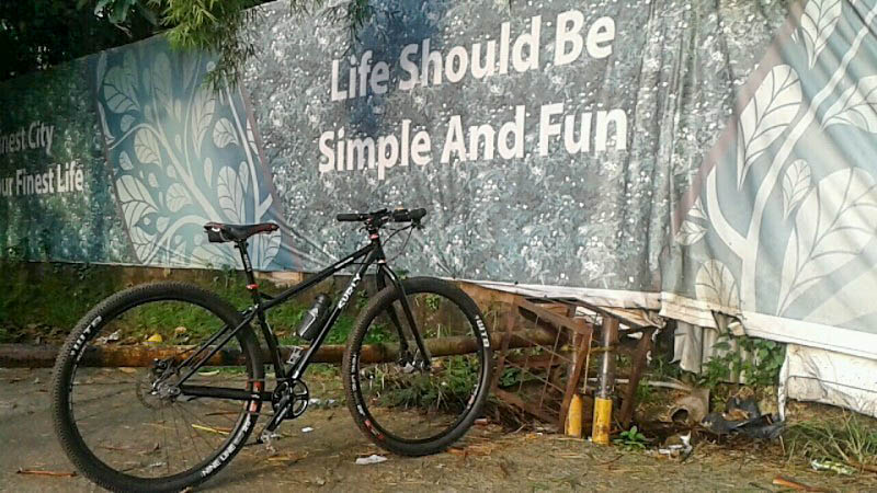 Right side view of a black Surly bike, parked on a dirt lot, facing a banner that reads, Life Should Be Simple And Fun