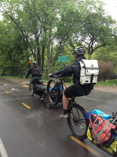 Rear, left side view of a cyclist, riding a wheelie on a Surly bike with a trailer, on a paved trail with trees behind