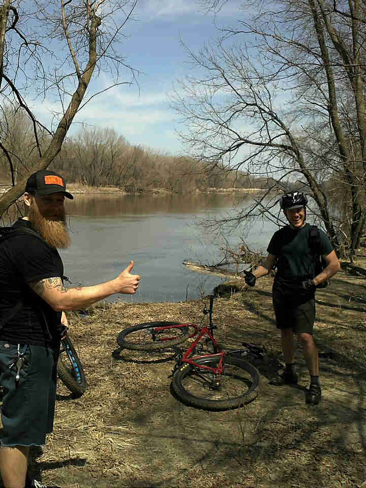 Two people showing thumbs up, standing on a dirt river bank with bare trees, and a red Surly bike laying between them