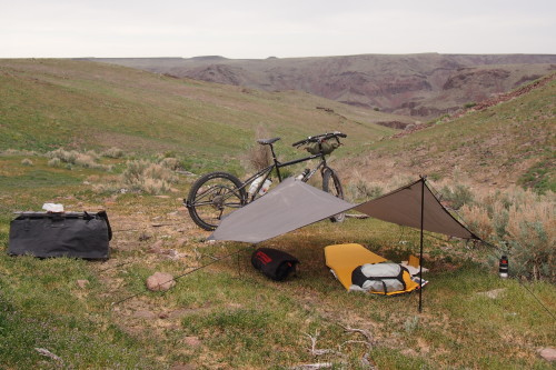 Right side view of a Surly Big Dummy bike, behind a tarp tent with a sleeping bag under it, on a hilly grassy field