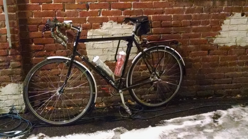 Left side view of a black bike, with 2 water bottles, parked against a red brick wall