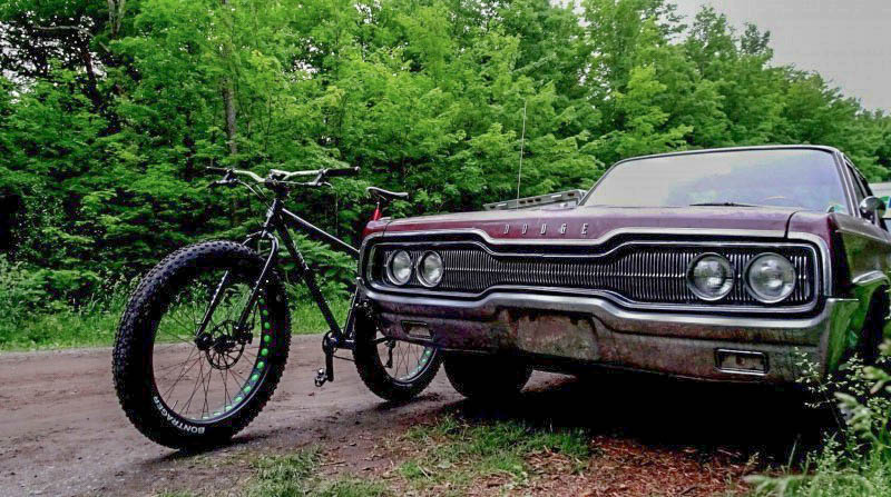 Left side view of a Surly fat bike peeking out from the front left side of an old Dodge car on the side of a gravel road