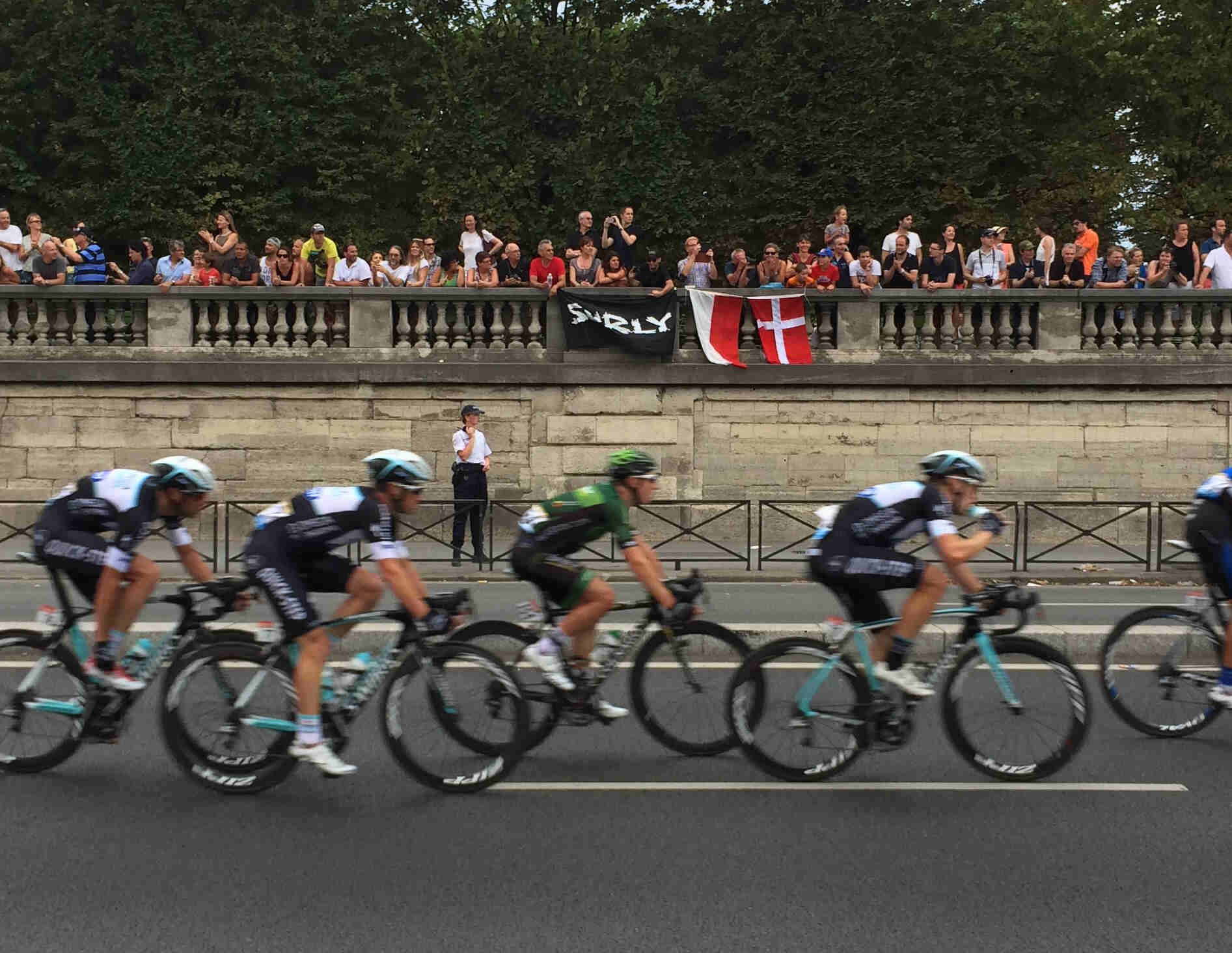 Right side view of cyclists riding their bikes down a street, with spectators above a wall, holding a Surly banner
