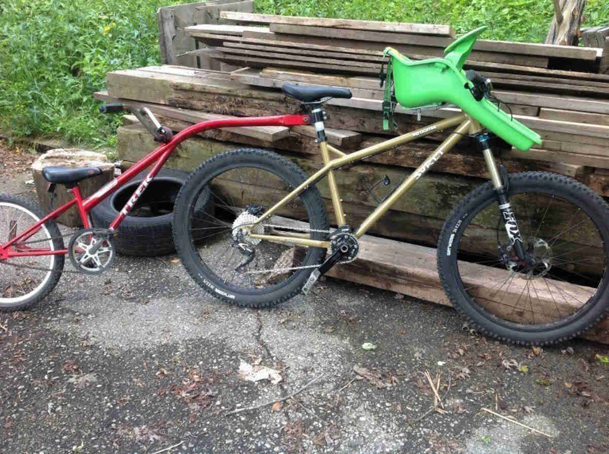 Right side view of a gold Surly Instigator bike with a handlebar child seat, and a child's trailer bike mounted behind