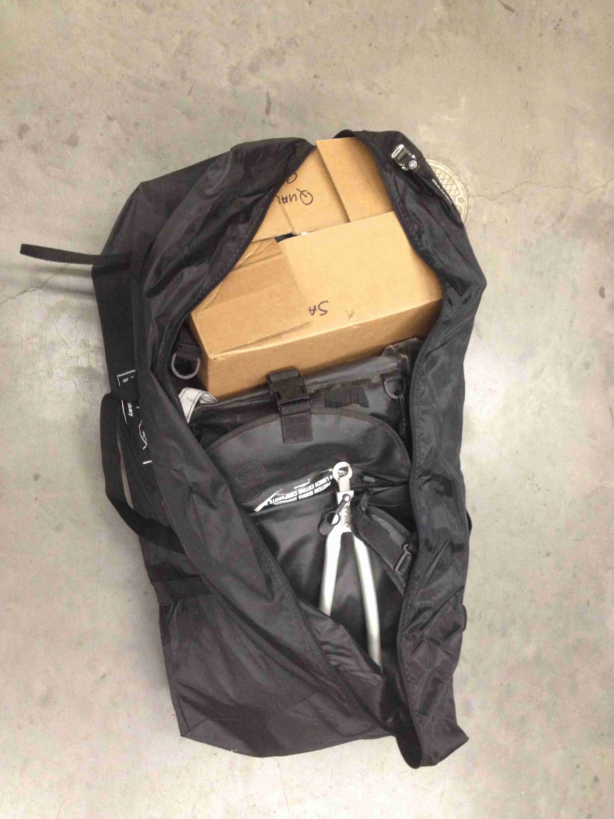 Downward view of an unzipped, black gear bag with a cardboard box and other items in it, on a cement floor