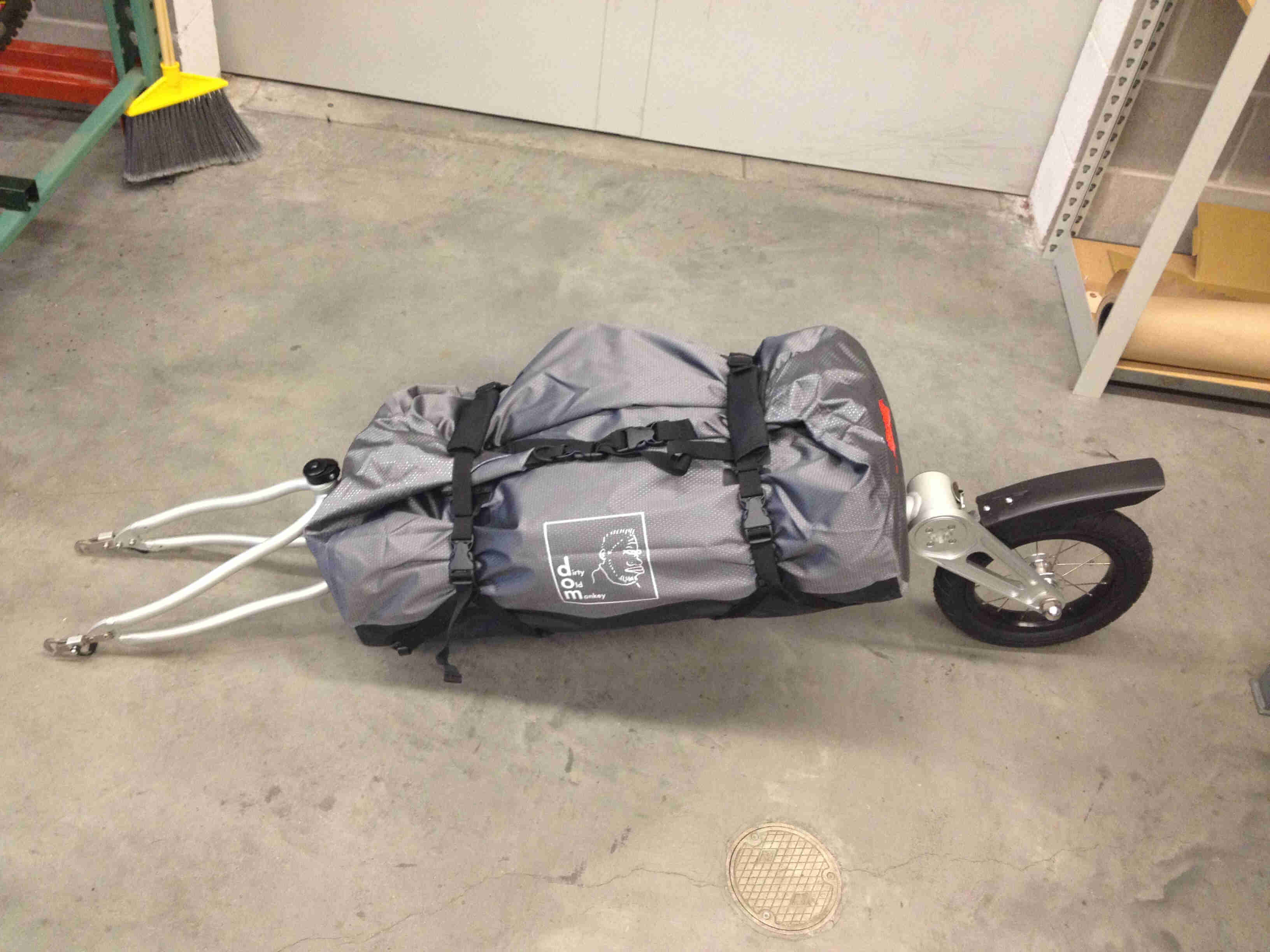 Downward, left side view of a foldable bike trailer, with a gray dry bag strapped to it, on a cement floor