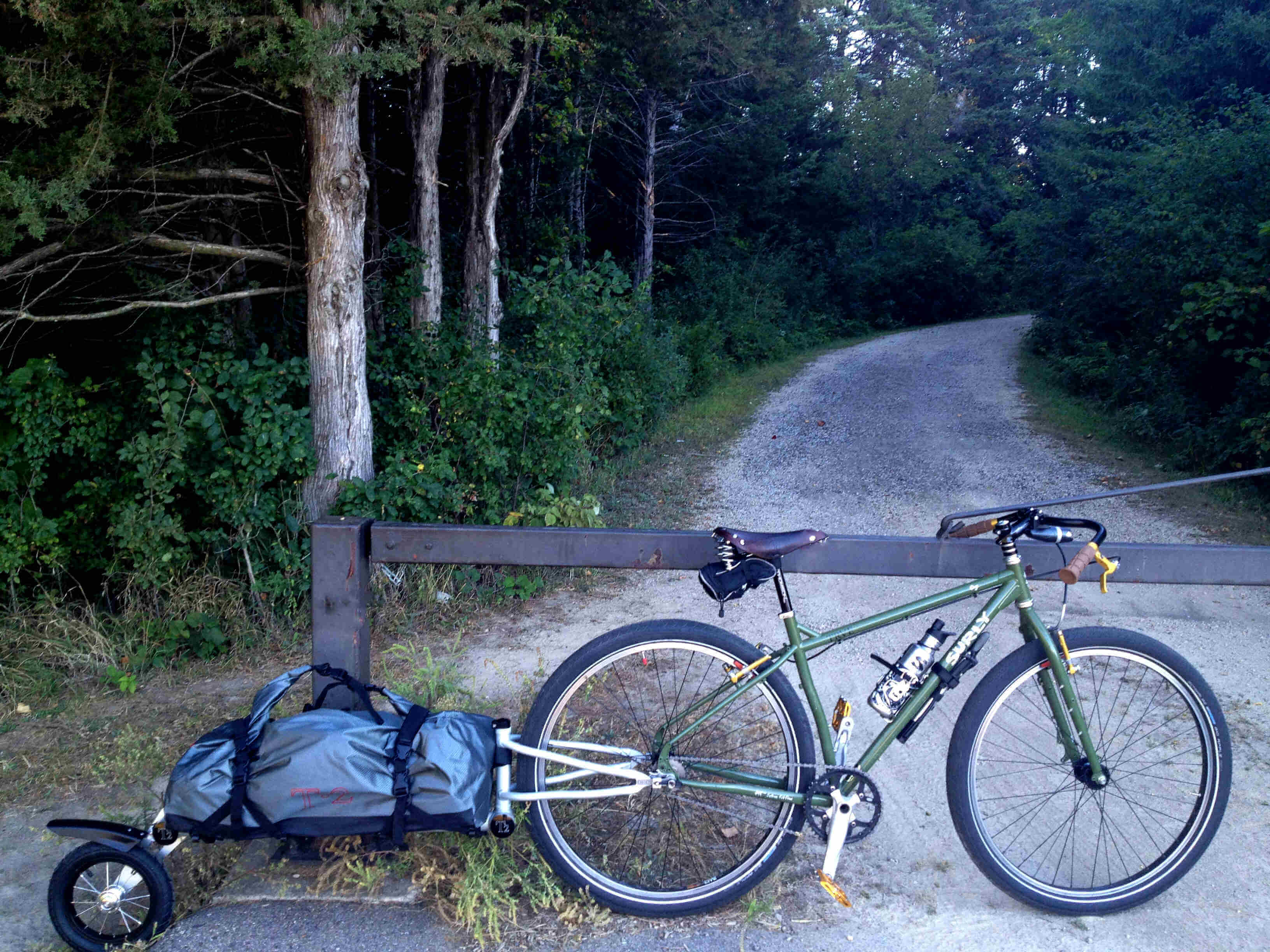 Right side view of a green Surly Ogre bike with foldable trailer, leaning on a gate with a gravel road behind it