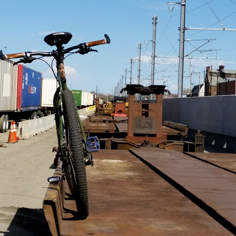 Rear view of a green bike, parked on the edge of a rusty, flatbed train car, in a rail yard 