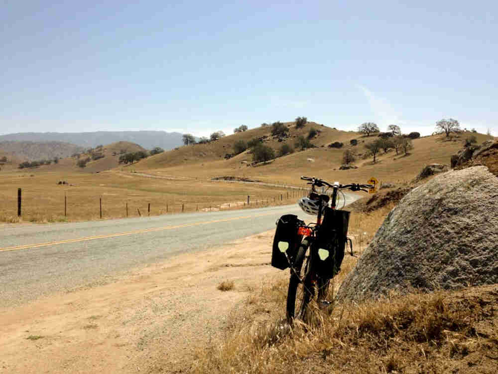 Rear view of a Surly Troll bike with gear, parked against a rock next to a paved road, facing a field and hills