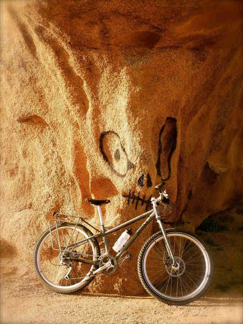 Right side view of tan Surly Troll bike, leaning on a sand cave wall with a face spray painted on it