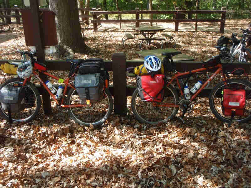 Side view of 2 orange Surly Troll bikes facing opposite directions, parked against a wood fence on leaf covered ground