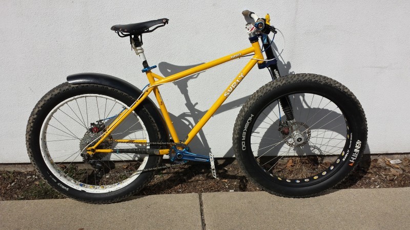 Right side view of a yellow Surly Pugsley bike, parked on a sidewalk, leaning against a white wall