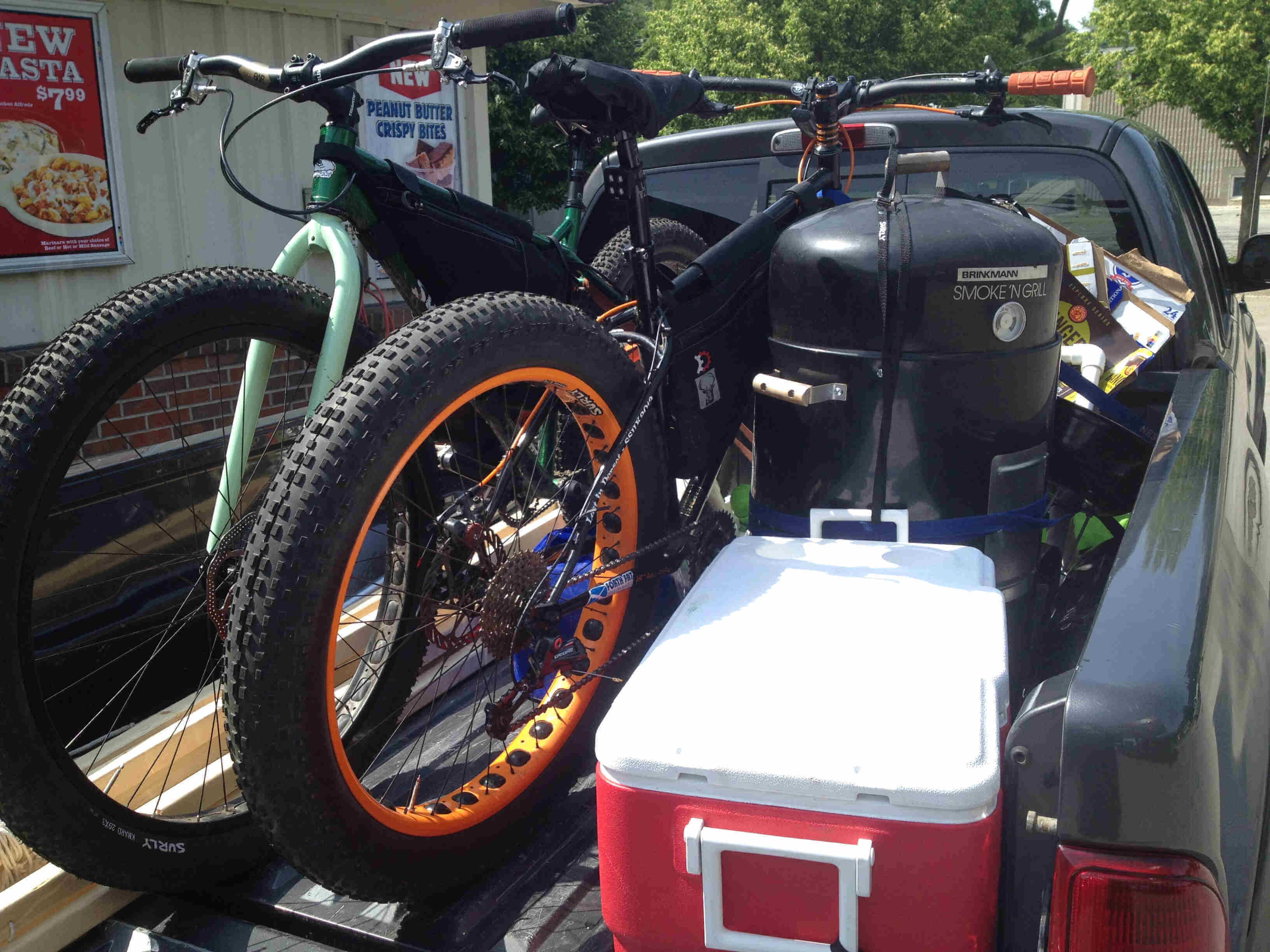 Rear, right side view of a 2 Surly bike, loaded into a pickup truck bed, with a food smoker and a cooler