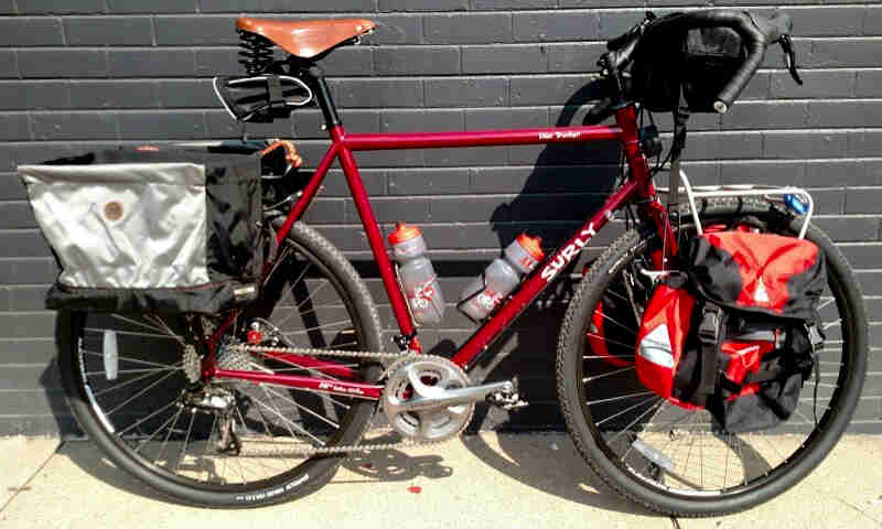 Right side view of a red Surly Disc Trucker bike, loaded with gear, parked on a sidewalk against a black brick wall